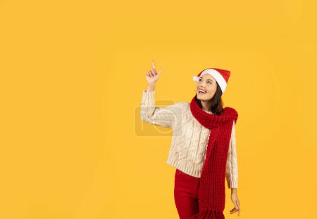 Photo for Cheerful lady in festive Santa hat pointing up to free space, perfect for holiday offers, set against vibrant yellow studio background, embodying excitement of winter holidays - Royalty Free Image