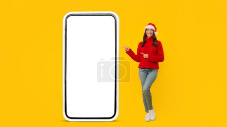 Photo for Smiling lady in festive knitwear presents big blank phone screen ready for Christmas offers, embodying the cheerful Xmas spirit of technology and communication in New Year. Panorama, mockup - Royalty Free Image