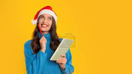 Photo for European woman smiles dreaming and writing down Christmas festive dreams and sales wishes in her notebook, standing on yellow studio background. Panorama, free space for text - Royalty Free Image