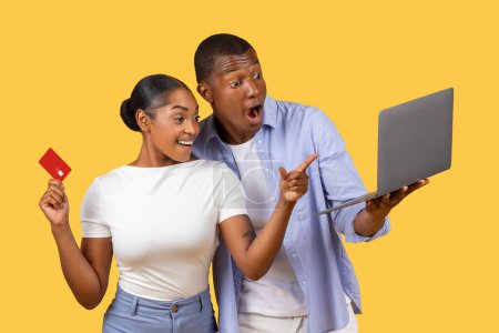 Téléchargez les photos : Les conjoints noirs excités tiennent l'ordinateur portable et la carte de crédit, la femme pointant avec enthousiasme vers l'écran, dans un contexte jaune vif, capturant l'ambiance vibrante des ventes saisonnières et des offres - en image libre de droit