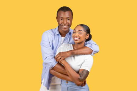 Radiant young black couple in loving embrace, sharing genuine smiles, dressed in smart casual wear standing on vivid yellow background