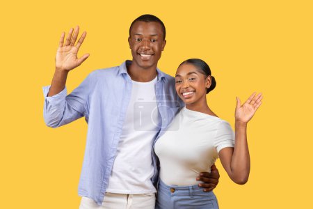 Téléchargez les photos : Couple noir souriant saluant avec une vague amicale, montrant les mains ouvertes sur un fond jaune joyeux, rayonnant de chaleur et souriant à la caméra - en image libre de droit