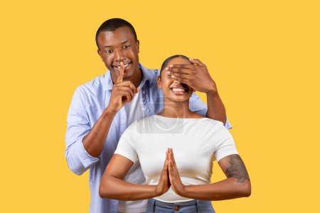 Téléchargez les photos : Homme noir souriant faisant un geste de silence tout en couvrant ludique les yeux de la femme en pose de prière, debout sur fond jaune vif - en image libre de droit