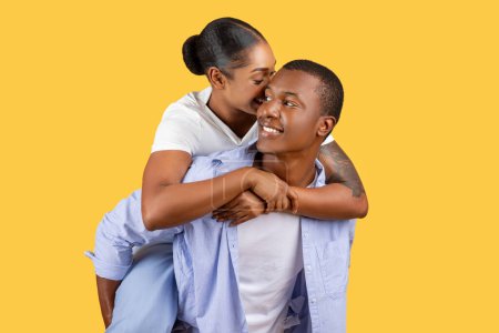 Téléchargez les photos : Femme noire affectueuse chuchotant dans l'oreille de ses partenaires, qui sourit avec un regard de contentement, tous deux habillés avec désinvolture sur fond jaune uni - en image libre de droit