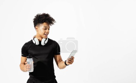 Photo for Brazilian guy checks fitness app on phone, ready with water bottle and earphones for workout on white studio backdrop, syncs music on smartphone with wireless headphones for training. Copy space - Royalty Free Image