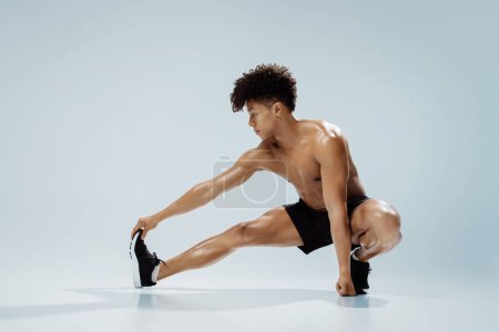 Photo for Fitness workout. Young shirtless athletic man exercising in extended leg squat pose, stretching legs and touching toes, demonstrating flexibility and strength over gray studio background - Royalty Free Image