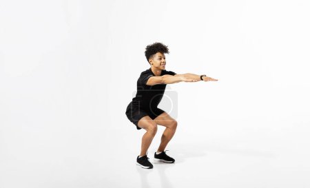 Foto de Hombre joven atlético que extiende los brazos mientras está en cuclillas durante el entrenamiento del gimnasio, trabajando en la fuerza de las piernas, usando ropa de deporte, haciendo ejercicio en el fondo del estudio blanco. Banner de fitness con espacio vacío - Imagen libre de derechos