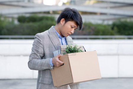 Photo for Korean businessman stands out of office building holds personal belongings in carton box outdoors, reflecting harsh reality of unemployment amidst corporate urban city backdrop - Royalty Free Image