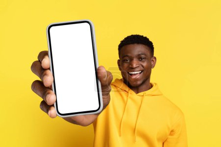 Foto de Feliz guapo sonriente joven negro en sudadera con capucha amarilla que muestra el teléfono grande con pantalla blanca en blanco, recomendando buena oferta en línea, aplicación móvil, aislado en el fondo amarillo, espacio de copia, maqueta - Imagen libre de derechos