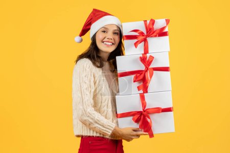 Téléchargez les photos : Joyeux dame tenant plusieurs boîtes de Noël empilent, posant gai et festif dans le chapeau de Père Noël, parfait pour les achats de vacances et les thèmes de célébration, studio tourné sur fond jaune - en image libre de droit