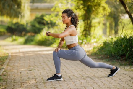 Photo for Young fitness woman in park lunging forward, checking heart rate on smartwatch, focused on weight loss and muscle tone, perfect for summer morning workouts and flexibility training outdoors - Royalty Free Image