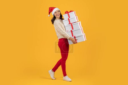Foto de Mujer alegre en el sombrero rojo de Santa Claus que lleva una pila de regalos de Navidad, expresando el estado de ánimo brillante de las vacaciones, caminando en el estudio en el fondo amarillo, ideal para el anuncio de invierno. Vista completa - Imagen libre de derechos