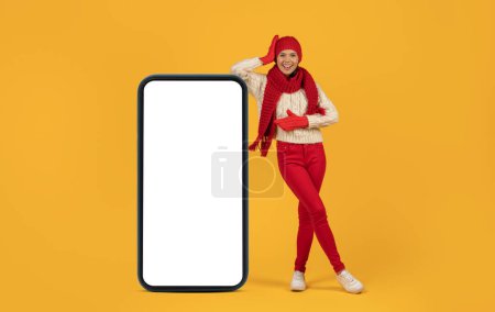 Téléchargez les photos : Joyeux jeune femme en chapeau rouge et écharpe, appuyé sur un téléphone énorme avec écran blanc, parfait pour l'application de vacances et des bannières de vente, studio tourné sur fond jaune. Maquette - en image libre de droit