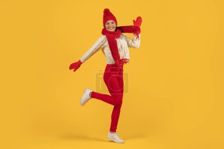Photo for Joyful woman in red knitted hat and scarf jumping mid air, full of New Year excitement, ideal for seasonal Xmas sale advertisements, studio shot against yellow background with free space - Royalty Free Image