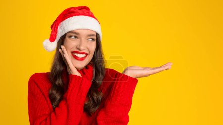 Photo for Joyful lady gestures with open hand palm on yellow studio background, wearing Santa hat. Christmas season best offers and holiday anticipation. Panorama, free space for text - Royalty Free Image