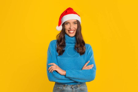 Téléchargez les photos : Joyeux jeune femme en pull décontracté et chapeau de Père Noël croise avec confiance ses bras, incarnant l'esprit de Noël festive, debout sur fond jaune vif en studio. Vente de Noël annonce - en image libre de droit