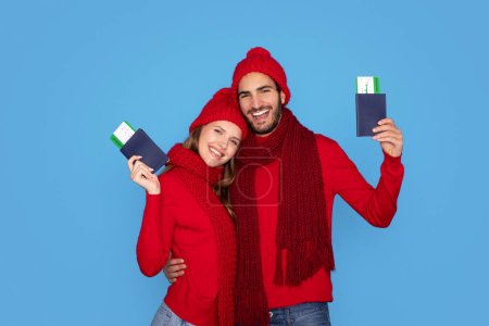 Photo for Travelling Concept. Young Smiling Couple Wearing Knitted Hats Holding Passports With Boarding Pass Tickets, Happy Man And Woman Ready For Winter Vacation, Posing Isolated On Blue Studio Background - Royalty Free Image