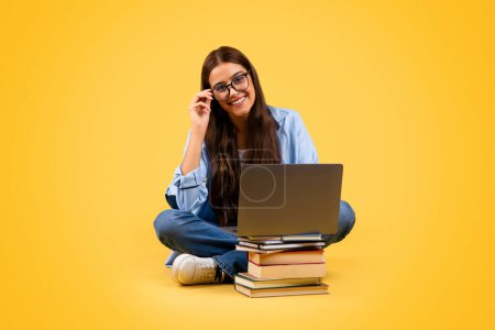 Téléchargez les photos : Heureuse adolescente européenne étudiante en lunettes utiliser un ordinateur avec des livres, dactylographier, étudier au sol, isolé sur fond jaune, studio, pleine longueur. Travail en ligne, éducation et leçon à distance - en image libre de droit