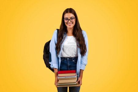 Positive Teenager-Studentin mit Brille hält viele Bücher in der Hand, genießt das Lernen und die Hausaufgaben, isoliert auf gelbem Studiohintergrund. Bildung, Lebensstil in der Schule, Werbung und Angebot an der Universität