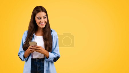 Téléchargez les photos : Heureuse étudiante caucasienne, tapant un message au téléphone, bavardant sur les réseaux sociaux, regardant l'espace libre, isolée sur fond jaune, panorama. Éducation et application d'étude en ligne - en image libre de droit