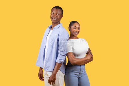 Téléchargez les photos : Couple afro-américain confiant debout dos à dos, souriant vivement en tenue décontractée sur fond jaune, femme aux bras croisés - en image libre de droit