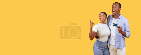 Téléchargez les photos : Jeunes conjoints noirs, tenant la carte de crédit, femme pointant énergiquement vers l'espace vide à côté d'eux sur fond de studio jaune vif, transmettant l'humeur du shopping pour les ventes saisonnières et les offres - en image libre de droit