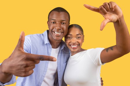 Téléchargez les photos : Couple noir souriant faisant ludique cadre de la caméra avec leurs mains, capturant moment de joie, dans des vêtements décontractés sur fond jaune joyeux - en image libre de droit