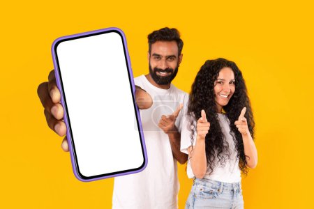 Téléchargez les photos : Application mobile. Heureux couple arabe montrant un énorme smartphone avec écran blanc et pointant vers la caméra debout sur fond jaune, maquette. Homme et femme excités montrant grand téléphone, modèle - en image libre de droit