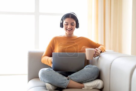 Photo for Young Indian Woman Wearing Wireless Headphones Resting With Laptop And Coffee On Couch, Happy Smiling Eastern Lady Using Computer At Home, Working Online Or Surfing Internet, Enjoying Hot Drink - Royalty Free Image