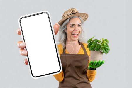Foto de Mujer agricultora alegre muestra enorme teléfono móvil con pantalla vacía, sosteniendo planta verde en maceta sobre fondo gris estudio. Aplicación de mercado de comestibles. Mockup, collage con el teléfono celular grande - Imagen libre de derechos