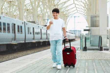 Téléchargez les photos : Asiatique en tenue décontractée touriste marchant par la gare, portant des bagages rouges, en utilisant un smartphone, vérifier l'horaire du train, acheter des billets en ligne, faire une réservation de taxi sur l'application mobile, copier l'espace - en image libre de droit