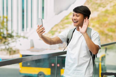 Téléchargez les photos : Gars asiatiques positifs avec sac à dos blogueur de voyage prendre selfie par téléphone, enregistrement vidéo pour les abonnés tout en se tenant sur la plate-forme à la gare, saluant à l'écran du smartphone, copier l'espace. Tourisme - en image libre de droit