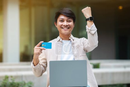 Photo for Emotional young asian man in casual clothing use laptop pc computer, hold credit bank card, shopping or banking online, raising hand up, clenching fist, copy space - Royalty Free Image
