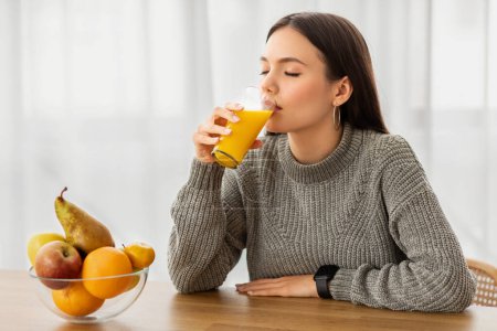 Photo for Joyful young brunette woman in casual clothes with long hair sitting at desk and drinking fresh orange juice near window, copy space. Healthy lifestyle, diet - Royalty Free Image