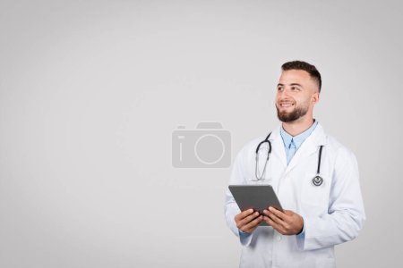 Photo for European young male doctor in white coat holding digital tablet, gazing at copy space over simple grey background, exemplifying modern healthcare - Royalty Free Image