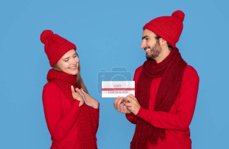 Feliz joven mujer recibiendo un certificado de regalo de su amado novio, alegre pareja joven con sombreros de punto y bufandas saludándose mutuamente con vacaciones de invierno, de pie sobre fondo azul