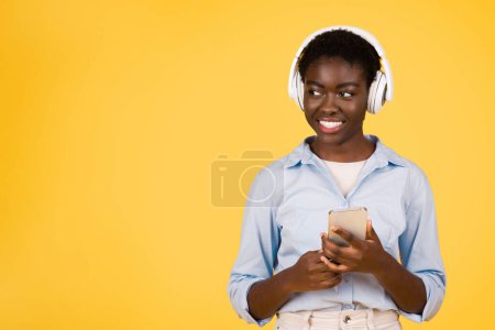 Photo for Smiling young african american woman student in wireless headphones, listen music, typing on smartphone, enjoy online lesson, isolated on yellow studio background. Study and education, spare time - Royalty Free Image