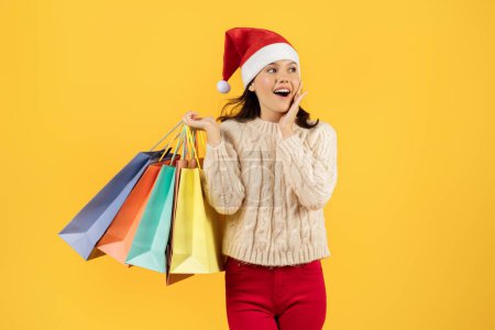 Photo for Happy young lady shopper in Santa hat and red knitwear, holding shopping bags full of holiday purchases, expressing excitement, perfect for seasonal sale ads, studio shot against yellow background - Royalty Free Image
