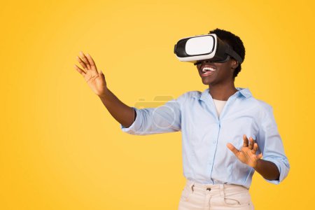Photo for Smiling young african american woman student in casual and vr glasses, controls copy space with hands, enjoy game, isolated on yellow studio background. Study and education, virtual reality - Royalty Free Image