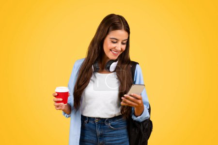Téléchargez les photos : Joyeux adolescent étudiante femme avec téléphone mobile, bavarder, profiter d'une tasse de café à emporter, isolé sur fond jaune. Temps libre avec application en ligne, pause d'étude et réseaux sociaux, musique - en image libre de droit