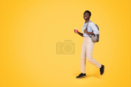 Téléchargez les photos : Heureuse jeune étudiante afro-américaine décontractée avec sac à dos marche avec tasse de café à emporter, profiter de temps libre, isolée sur fond de studio jaune. Études et éducation - en image libre de droit
