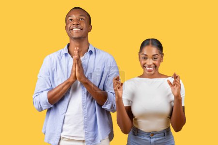 Photo for Black man praying with hands clasped and woman with crossed fingers smiling, both hoping for the best standing on yellow background - Royalty Free Image