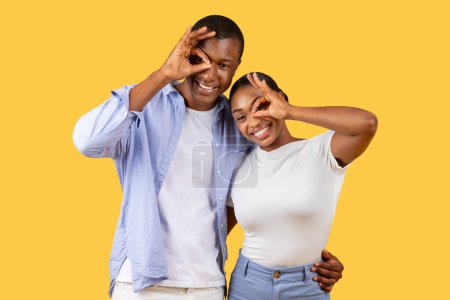 Téléchargez les photos : Souriant jeune couple noir ludique faire des lunettes de main, debout ensemble sur un fond jaune vibrant et souriant ensemble à la caméra - en image libre de droit