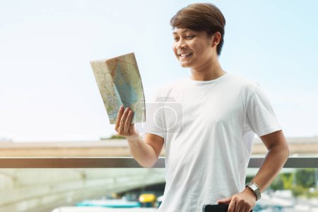Foto de Primer plano de alegre joven asiático hombre turista llegó a destino de viaje. Posando al aire libre junto al aeropuerto con el equipaje, mirando el mapa de papel y sonriendo, disfrutando de sus vacaciones, espacio para copiar - Imagen libre de derechos