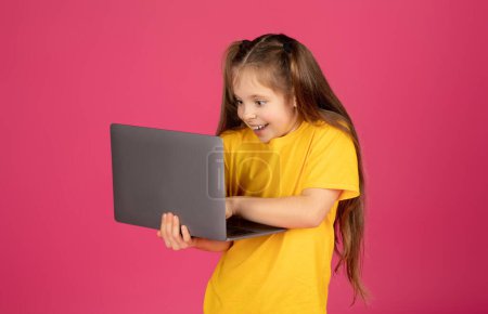 Téléchargez les photos : Petite fille excitée jouant à des jeux en ligne sur ordinateur portable, Enfant préadolescent gai regardant l'écran d'ordinateur, Profitant d'un divertissement virtuel, Debout sur fond rose, Espace de copie - en image libre de droit