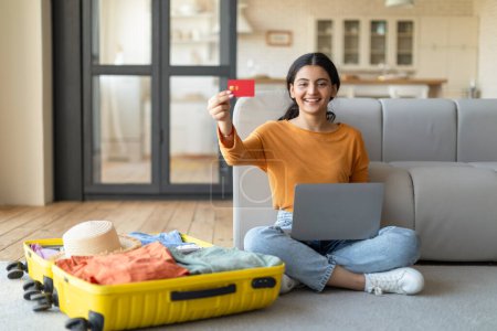 Photo for Happy young indian woman with credit card and laptop booking vacation travel, smiling eastern female sitting on floor next to suitcase with clothes, preparing for trip, recommending easy payments - Royalty Free Image