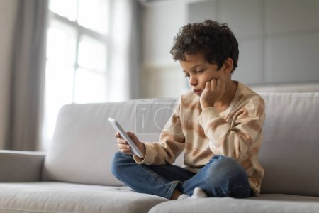 Foto de Aburrido niño preadolescente negro sentado en el sofá y el uso de teléfono inteligente en casa, molesto niño afroamericano masculino mirando a la pantalla del teléfono móvil, jugando juego en línea, sentirse solo, espacio de copia - Imagen libre de derechos