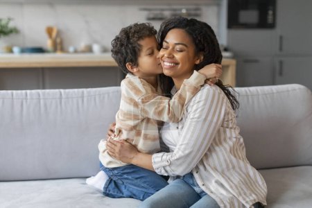 Foto de Preteen negro chico besos su feliz madre mientras ellos vinculación en casa, amar afro-americano familia mamá y macho niño abrazando y sonriendo mientras sentado en sofá en sala de estar interior, copiar espacio - Imagen libre de derechos
