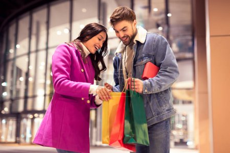 Photo for Cheerful buyers couple on Xmas gifts shopping looking inside bags, husband holds present box, standing together outside modern mall in the city at night. Seasonal buying and family surprises - Royalty Free Image