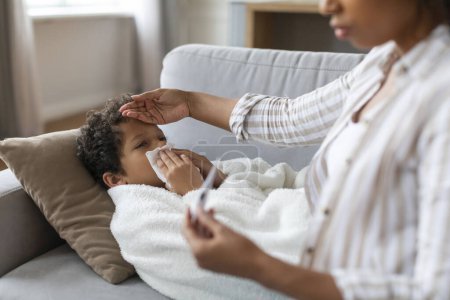 Photo for Seasonal Flu Concept. Worried Black Mom Checking Temperature Of Her Ill Son At Home, Caring Mother Touching Childs Forehead And Looking At Thermometer, Taking Care About Sick Kid, Closeup Shot - Royalty Free Image